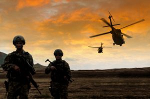 Two soldiers walking in the battlefield while two military helicopter flying over them during a military operation at sunset.